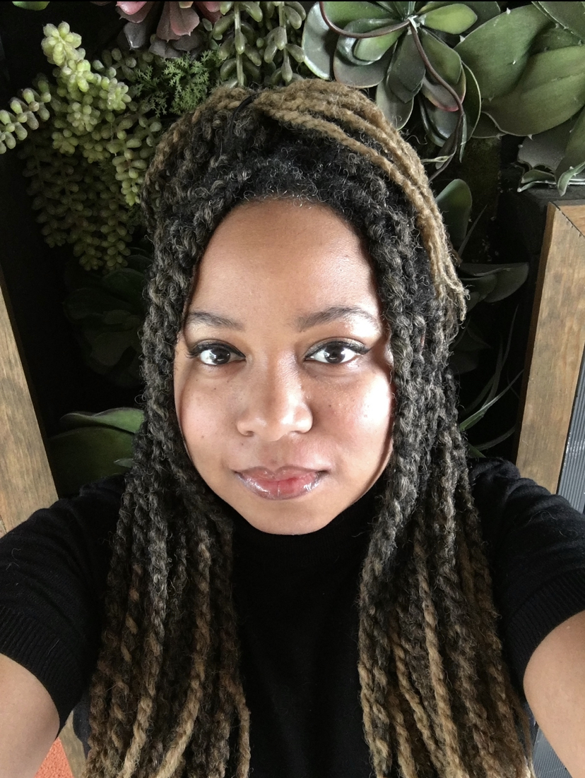 Erin Brown. A young black women with her hair in long black twists that are blonde at the tips, wearing a black short sleeve sweater in front of a wall of green plants.